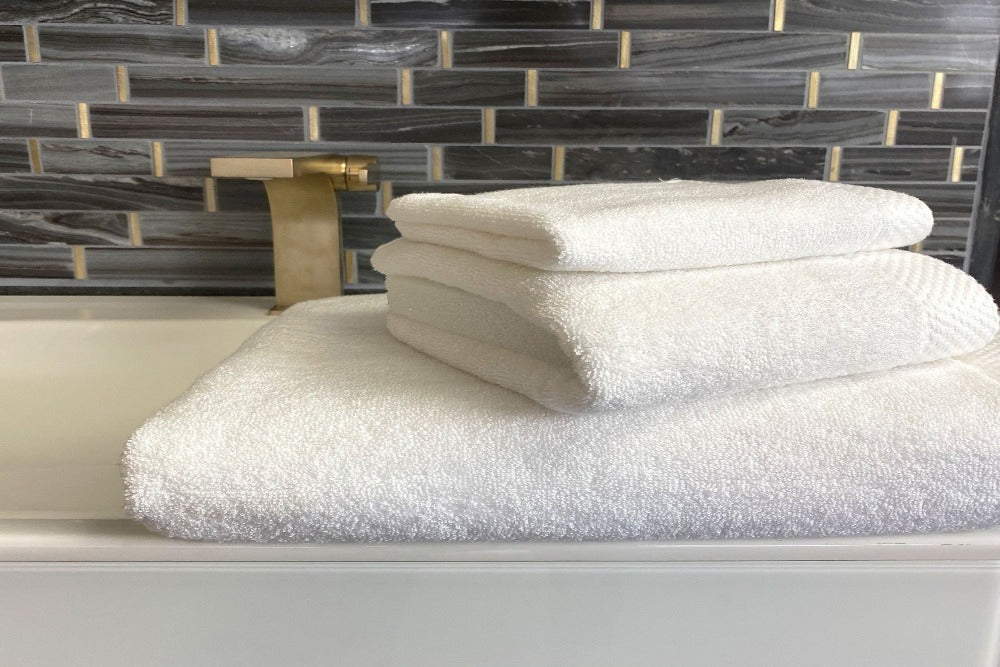 three neatly folded White Colored Luxurious Bamboo Cotton Bath Towels stacked by size and placed on the edge of a modern sink in a well decorated bathroom