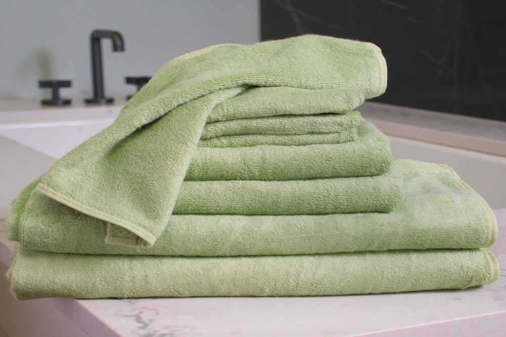 An 8 piece set of Sage Colored Luxurious Bamboo Cotton Bath Towels stacked by size and placed on a modern well decorated vanity