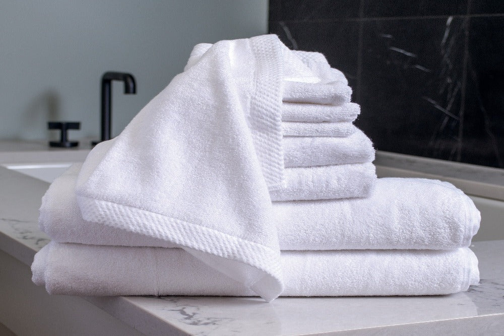 An 8 piece set of White Colored Luxurious Bamboo Cotton Bath Towels stacked by size and placed on a modern well decorated vanity