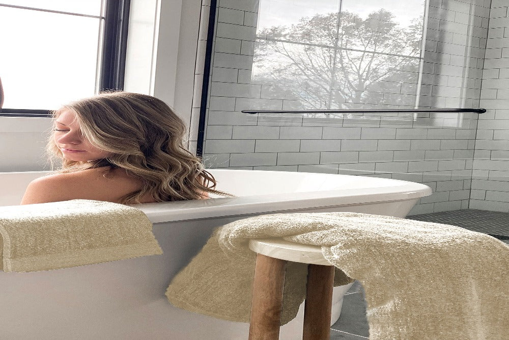 a back head and shoulder view of a peaceful attractive blond woman in a claw bathtub with her Champagne Colored Luxurious Bamboo Cotton bath towel and matching hand towel draped over the bathtub and a matching towel draped over a stool near the tub