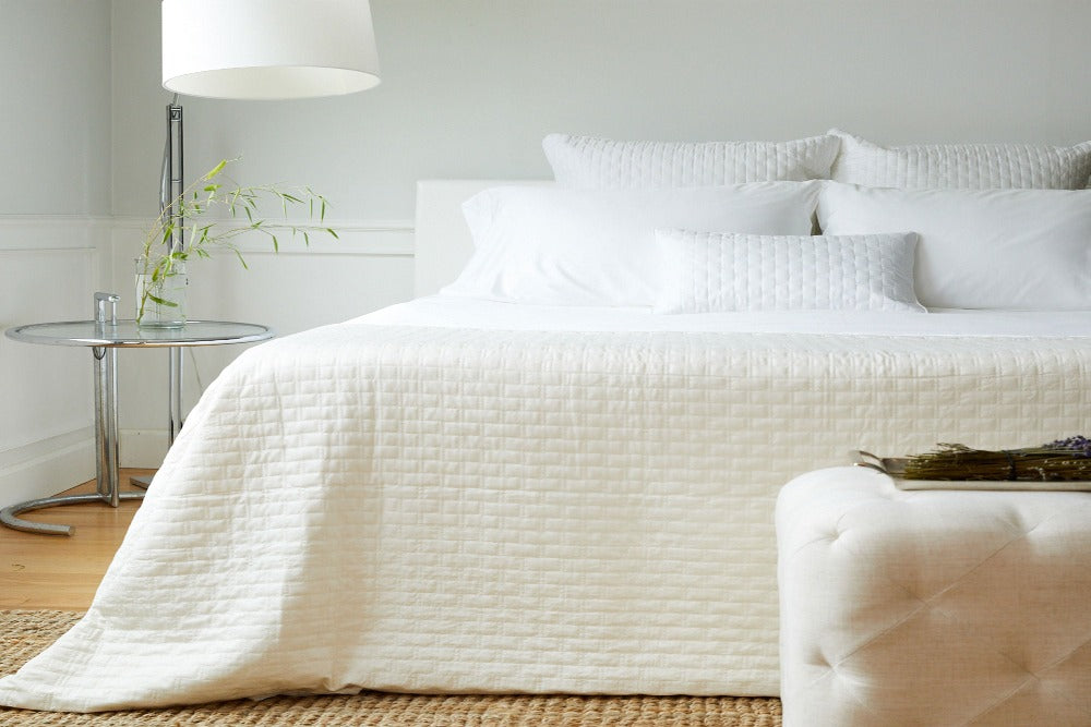 View of a bed in a light colored well decorated bedroom with a White Colored Luxurious 100% Bamboo Quilted Coverlet draped over the smartly made bed and five matching pillows at the headboard